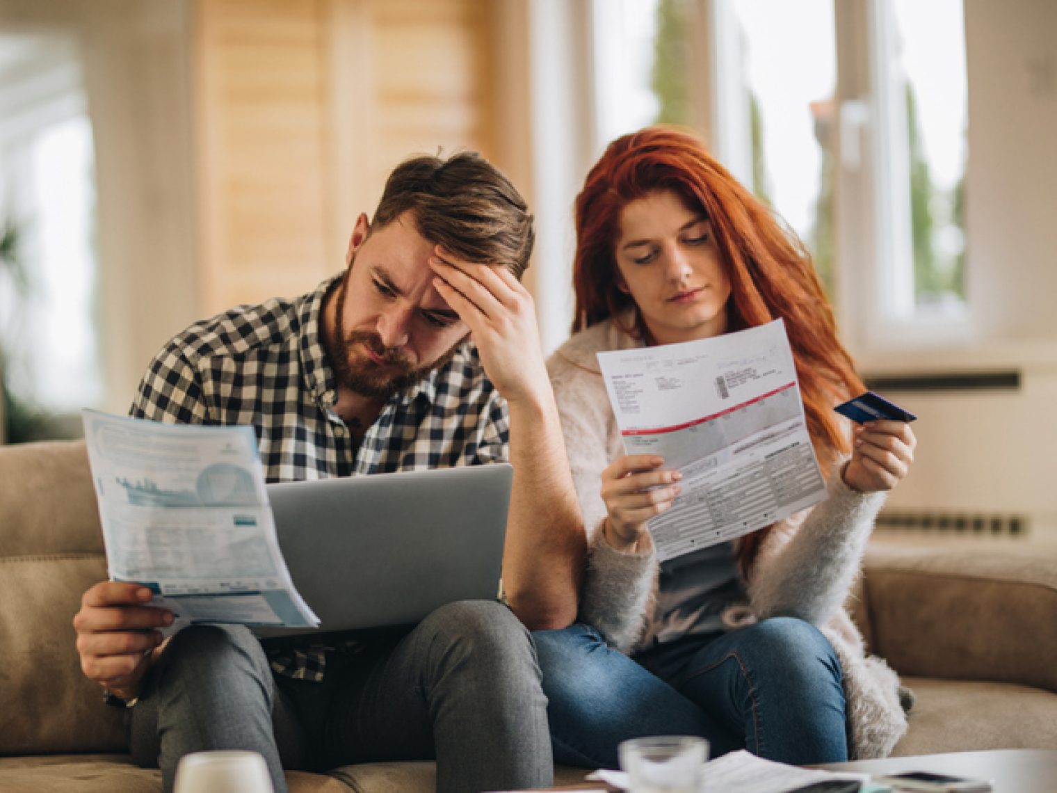 A worried couple is looking at their finances with a sense of concern. The man is holding a document and touching his forehead while the woman is holding another document and a credit card.