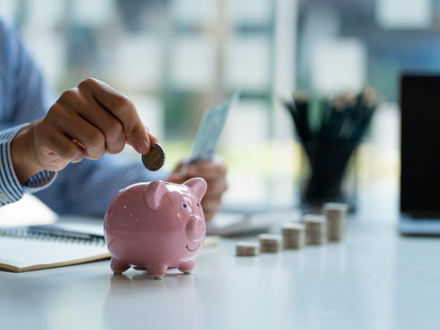 A person in a striped shirt is inserting a coin into a pink piggy bank, with a laptop and stacks of coins on a desk, symbolizing savings or financial planning.