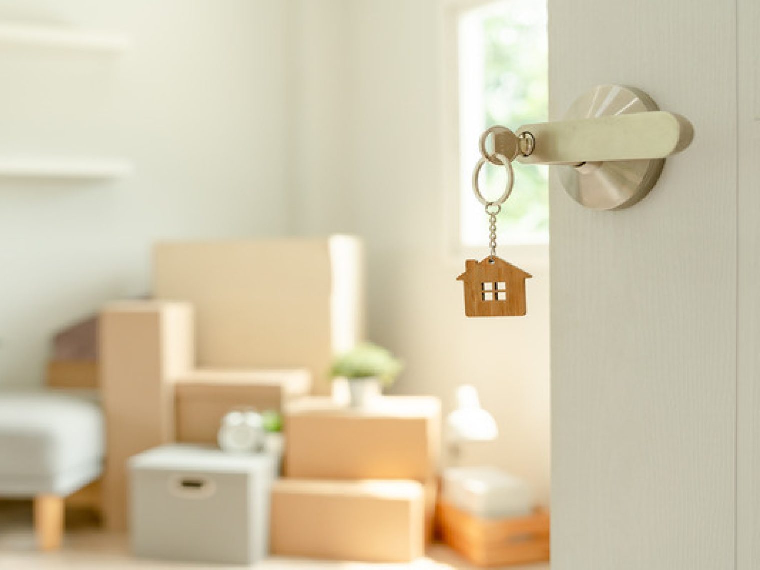 A pair of house keys with a house-shaped keychain inserted into the lock of an open door, with a bright, airy room in the background featuring cardboard moving boxes and minimalist furniture, symbolizing new homeownership.