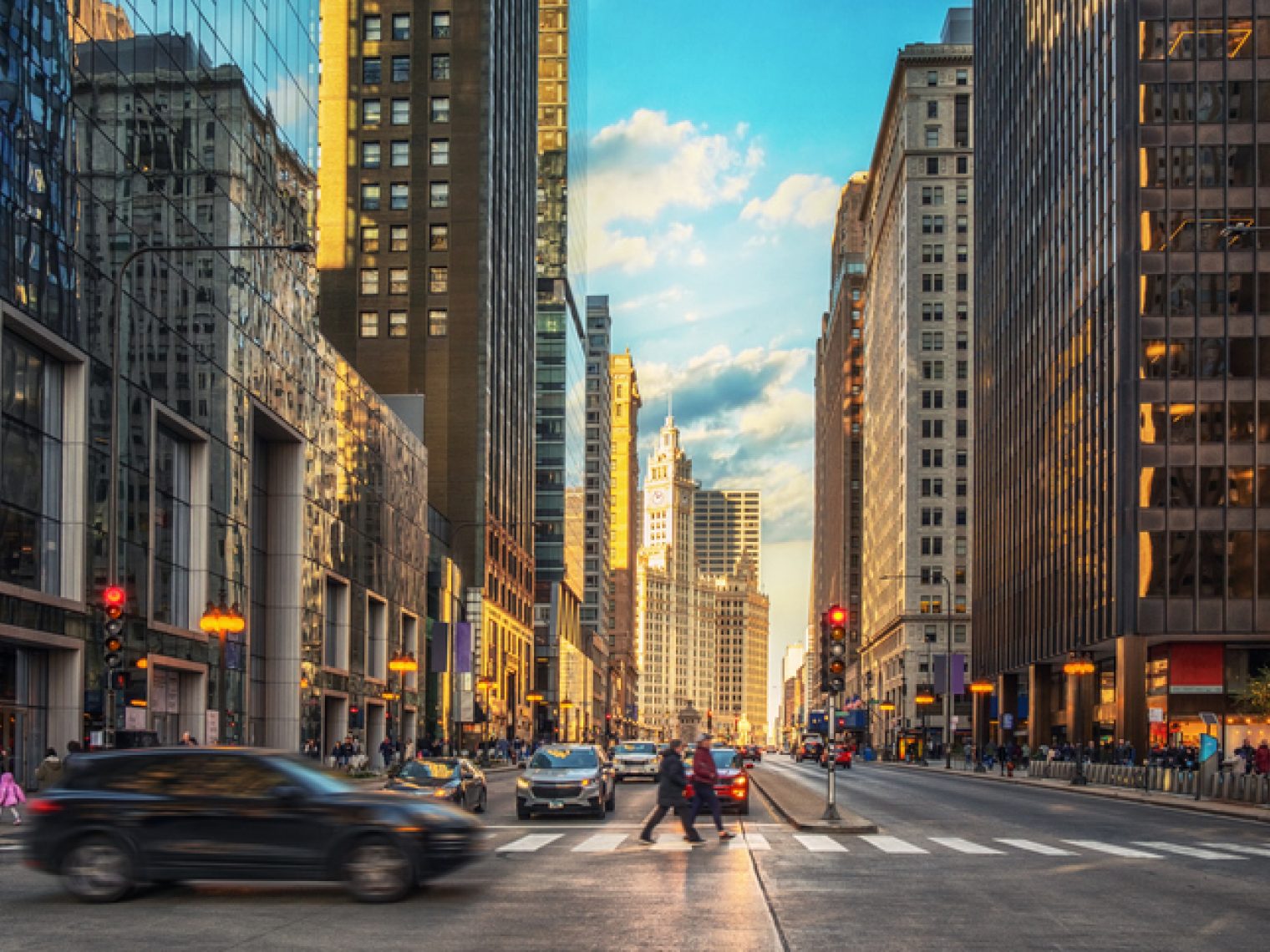 A bustling city street scene with cars in motion and pedestrians crossing, flanked by towering skyscrapers under a sky showing the transition from sunset to evening.