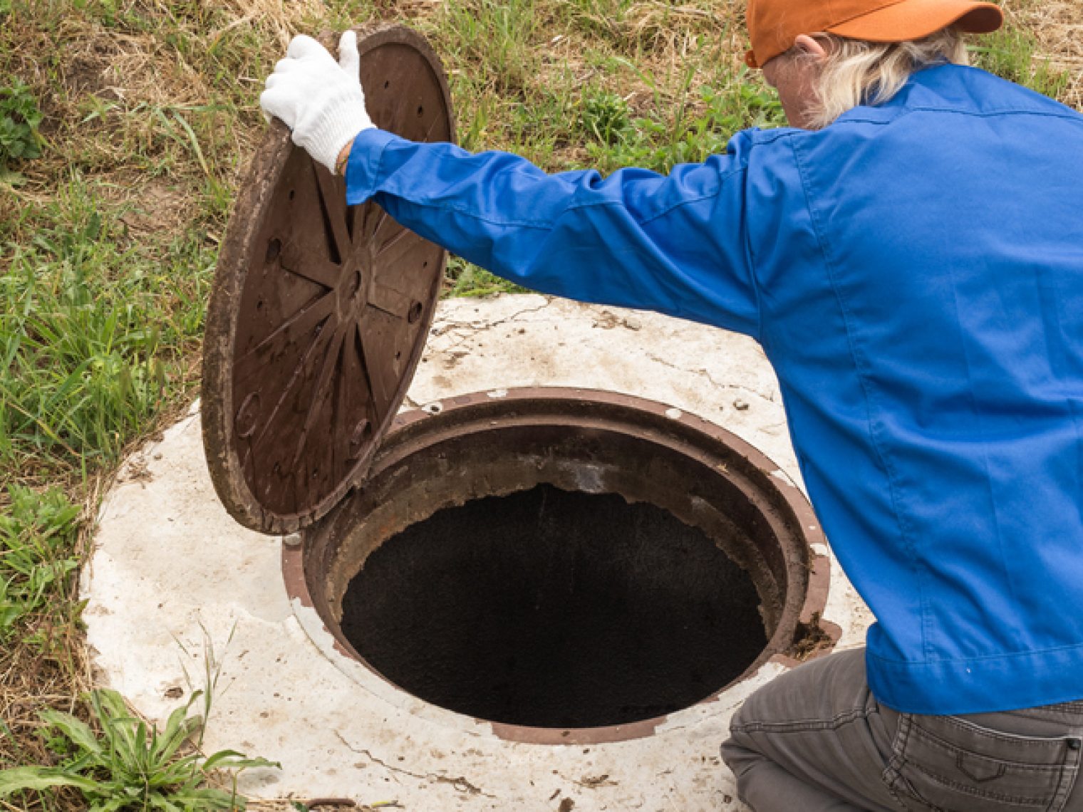 A worker wearing gloves and a blue jacket is opening a large, round sewer manhole cover on a concrete surface with grass around it.