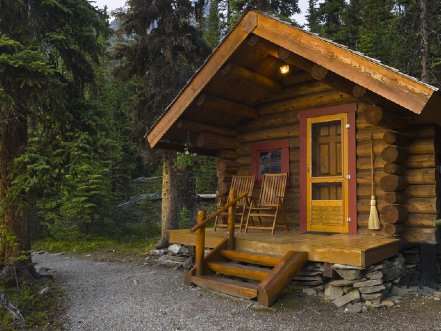 Nestled in a lush coniferous forest, this charming log cabin invites tranquility with its rustic design and natural surroundings. The cabin features a small porch with wooden steps leading to an orange door, complemented by two matching chairs that offer a spot to enjoy the serene environment.